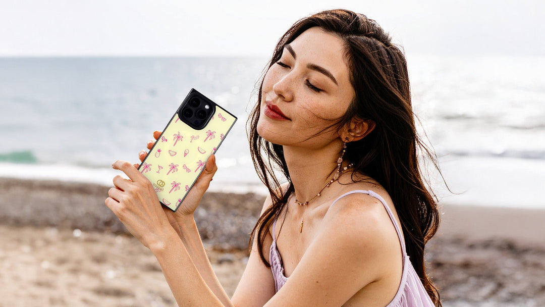 Woman at the beach holds a pale yellow phone case with a pink palm tree print