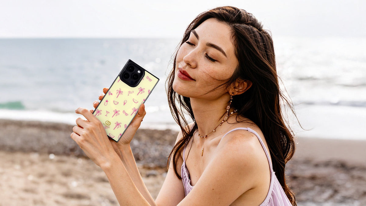 Woman at the beach holds a pale yellow phone case with a pink palm tree print
