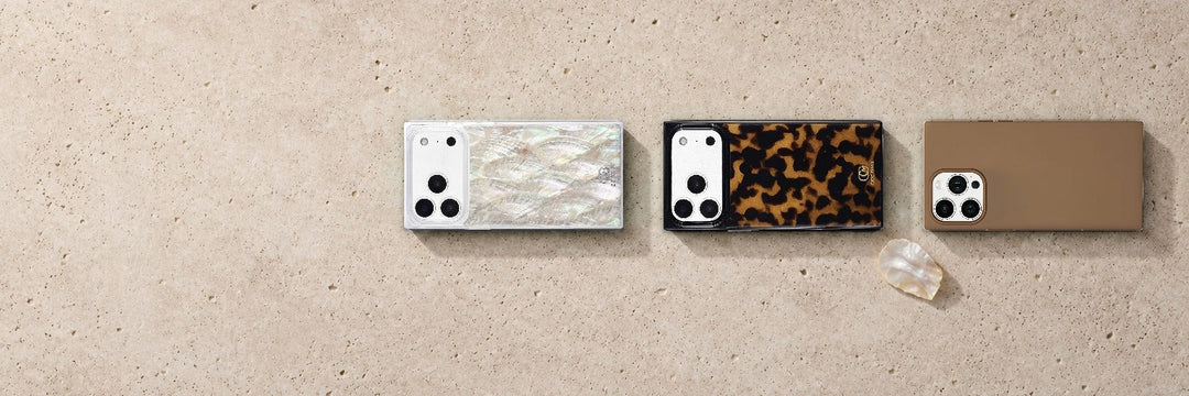 Three COCOMII rectangle iPhone cases in Mother of Pearl, tortoiseshell print and tan lying in a row on pale limestone beside a shell fragment