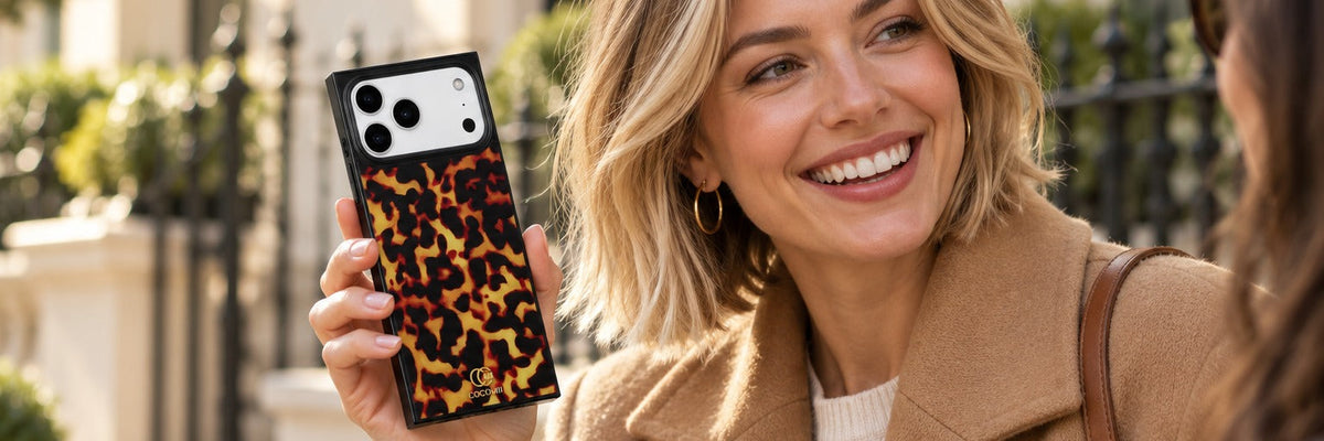 Woman in a camel coat with gold bangles holding a COCOMII tortoiseshell pattern case in Amber beside her face