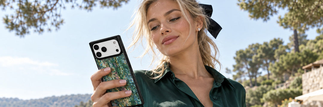 Woman in an emerald silk shirt dress on a hotel jetty glancing at her phone with a COCOMII abalone finish case in Aurora